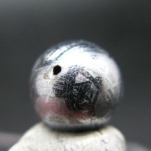 May include: A small, round, silver-coloured meteorite with a hole in the centre. The surface of the meteorite is covered in a pattern of lines and grooves.