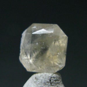 Könnte beinhalten: Ein durchscheinender, blass champagnerfarbener Edelstein mit einem facettierten, mehrseitigen geometrischen Schliff. Der Kristall zeigt innere Klarheit und lichtreflektierende Oberflächen und ruht auf einer strukturierten, hellgrauen, unregelmäßigen Steinbasis.