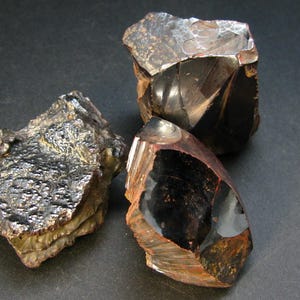 Könnte beinhalten: Drei Stücke schwarzer Hämatit mit irisierenden Oberflächen. Hämatit ist ein dunkles, metallisches Mineral mit einer glänzenden, reflektierenden Oberfläche.