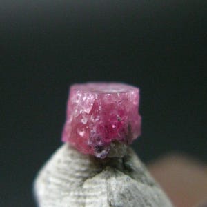 May include: A small, raw, cylindrical-shaped gemstone with a vibrant magenta-pink hue. The crystal features a rough, multifaceted, and textured surface with visible crystalline structures throughout its body, displaying deep pink and translucent tones.