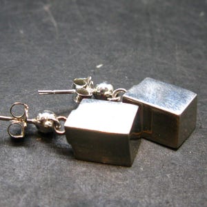 May include: A pair of silver cube stud earrings. Each earring features a small, polished silver cube suspended from a post with a butterfly clasp. The earrings are displayed on a dark, textured surface.