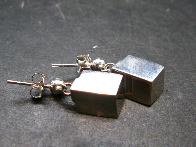 May include: A pair of silver cube stud earrings. Each earring features a small, polished silver cube suspended from a post with a butterfly clasp. The earrings are displayed on a dark, textured surface.