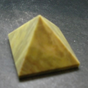 May include: A small, four-sided pyramid object with a smooth, matte surface. The pyramid features a muted, earthy yellow-green colour with subtle, darker mottled patterns throughout its structure, creating a consistent and uniform appearance.