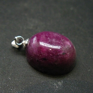 Könnte beinhalten: Ein silberner Anhänger mit einem tiefvioletten Edelstein. Der Edelstein ist oval geformt und hat eine glatte, polierte Oberfläche.