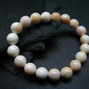Könnte beinhalten: Ein Armband aus runden, polierten Perlen in Weiß- und blasspfirsichfarbenen Tönen. Die Perlen sind aufgefädelt und bilden eine kreisförmige Form. Das Armband wird vor einem dunklen, strukturierten Hintergrund präsentiert.