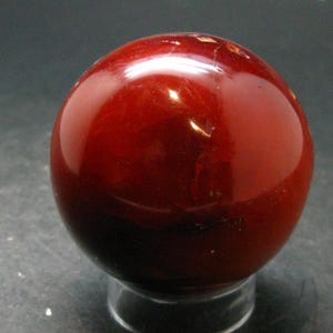 May include: A polished, spherical stone with a deep, rich red colour and a smooth, glossy surface. The sphere exhibits subtle internal variations and natural markings within its translucent, dark red material, resting on a small, clear base.
