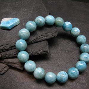 May include: A bracelet featuring round, light blue larimar beads. A small, irregularly shaped larimar stone is also visible. The jewellery is presented on a dark, textured surface.