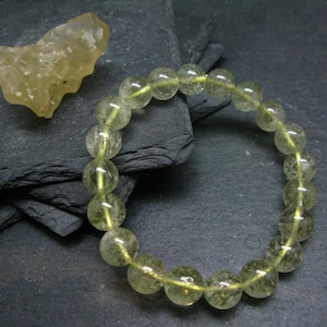 Może przedstawiać: Bransoletka wykonana z jasnozielonych, przezroczystych, okrągłych koralików. Koraliki są nawleczone na przezroczysty sznurek elastyczny. Bransoletka leży na ciemnoszarym tle.