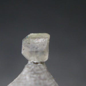 Könnte beinhalten: Nahaufnahme einer hellfarbigen, kristallinen Mineralprobe. Der Kristall hat eine geometrische Form mit leicht durchscheinendem Aussehen. Die Basis ist heller und der Hintergrund ist ein gedämpftes Grau.