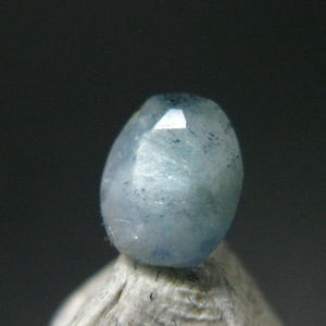 May include: A light blue gemstone with a faceted top and a speckled appearance. The oval-shaped stone is set against a neutral background, highlighting its color and texture. The gem is likely a bead or a component for jewelry making.