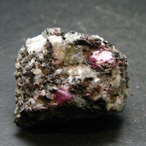 Peut inclure: Une pierre précieuse brute, non taillée, présentant un mélange de couleurs. La roche est composée de minéraux gris, noirs et blancs, avec plusieurs cristaux roses et rouges vifs incrustés. La texture est rugueuse et irrégulière.