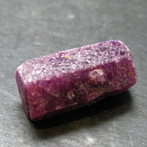 May include: A raw, uncut ruby gemstone. The ruby is a deep, rich magenta color with a rough, textured surface. The gemstone is rectangular in shape and appears to be natural and unpolished. The ruby is set against a dark gray background.