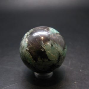 Puede incluir: Una esfera de piedra preciosa pulida con una mezcla de colores verde oscuro y marrón. La esfera tiene un acabado brillante y se apoya en un pequeño soporte transparente. La superficie de la piedra tiene un aspecto veteado.