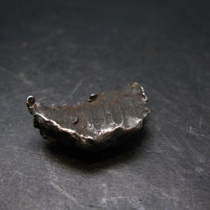 May include: A small, irregularly shaped, silver-toned metal object with a textured surface. The object has a curved form with a small loop at one end. The overall appearance suggests a handcrafted or artisan-made piece.