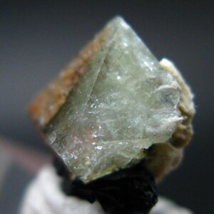 Puede incluir: Primer plano de un cristal translúcido verde claro con forma geométrica. El cristal tiene una textura ligeramente rugosa y está parcialmente encerrado en una matriz beige. La base del cristal es oscura.