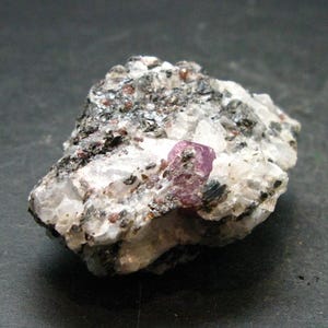 May include: A natural mineral specimen with a prominent pink crystal set within a matrix of white, grey, and black rock. The stone displays a rough texture, with visible crystalline structures and small, dark inclusions.