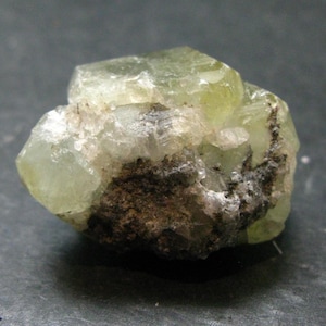 May include: A raw, translucent pale green crystal specimen with multifaceted surfaces and visible internal inclusions. The mineral features a cluster of angular, light-colored crystalline structures contrasted by a dark, textured earthy matrix.