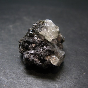 Puede incluir: Primer plano de un espécimen mineral con una matriz oscura y rugosa y cristales incrustados. Los cristales son translúcidos y de color claro, contrastando con el material base más oscuro y texturizado.