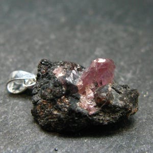 Puede incluir: Un colgante de plata con un grupo de cristales rosas y transparentes engastados en una matriz de roca volcánica negra.