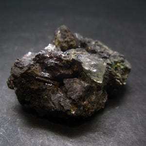 Puede incluir: Primer plano de un espécimen mineral oscuro y rugoso. La roca es principalmente de color gris oscuro y negro, con estructuras cristalinas y superficies reflectantes. Se ven pequeñas formaciones cristalinas claras, añadiendo contraste.