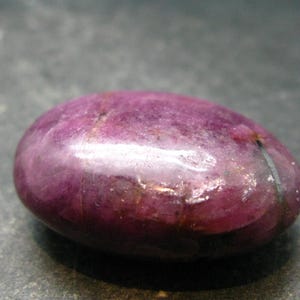 Könnte beinhalten: Ein polierter, oval geformter Rubin-Edelstein mit einem tiefen Magenta- und Violettton. Die Oberfläche des Steins weist natürliche Einschlüsse, subtile Variationen in der Farbintensität und eine glatte, reflektierende Textur auf.