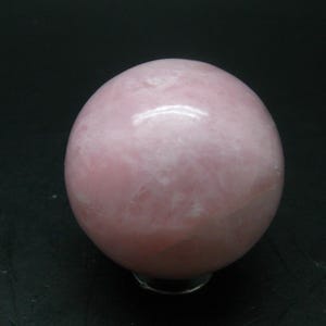May include: A smooth, spherical rose quartz crystal ball. The sphere is a pale pink colour with slight variations in tone and texture. The crystal ball is displayed on a clear stand against a dark background.