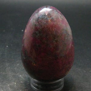 Könnte beinhalten: Ein polierter, eiförmiger Stein mit einer tief magentafarbenen und dunkelroten Grundfarbe, durchzogen von gesprenkelten schwarzen, grauen und metallisch goldfarbenen Einschlüssen auf der glatten, abgerundeten Oberfläche, auf einem kleinen, klaren Rundständer.
