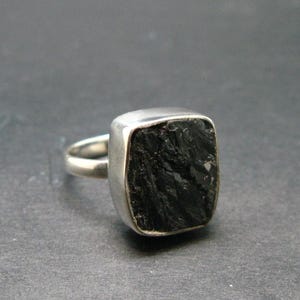 May include: A silver ring featuring a large, square-cut black gemstone. The stone has a textured surface and is set in a simple, polished silver band. The ring is displayed against a dark gray background.