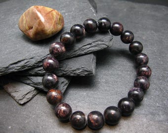 May include: A beaded bracelet with dark, polished, round beads, likely gemstones, in shades of black and burgundy. A smooth, oval-shaped stone with brown and tan patterns sits nearby. The bracelet is displayed on a dark, textured surface.