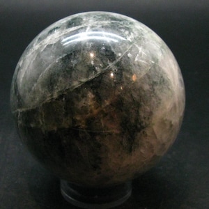 May include: A polished spherical stone featuring a marbled pattern of dark forest green, charcoal gray, and translucent white inclusions. The surface displays natural crystalline veins and reflective highlights across its smooth, rounded form.