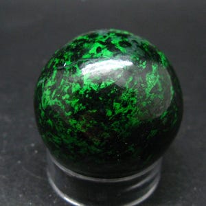 Puede incluir: Una piedra esférica pulida que presenta un patrón vibrante y moteado de verde bosque profundo y negro. La superficie es lisa y reflectante, mostrando una distribución orgánica e intrincada de inclusiones minerales verde claro y oscuro.