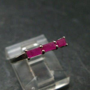 May include: A silver-toned ring featuring three rectangular, vibrant pink stones set in a row. The stones are held in place by small metal prongs, and the band is thin and metallic, showcasing a clean, linear design with a polished finish.