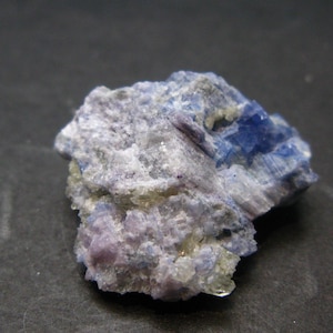 May include: A close-up of a rough, natural mineral specimen. The stone features a mix of colors, including shades of blue, lavender, and white. The texture is uneven, with visible crystalline structures. The overall shape is irregular.