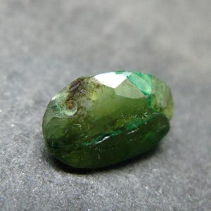 May include: A faceted oval gemstone in a deep forest green hue. The stone features a translucent quality with visible internal inclusions and natural variations in color intensity, displaying a polished surface with multiple geometric facets.