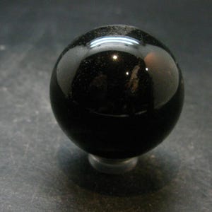 Puede incluir: Una esfera negra lisa y pulida con una superficie reflectante. El objeto es perfectamente redondo y de color oscuro, mostrando un reflejo de luz blanca brillante en su curva superior, lo que indica un acabado brillante y una composición sólida.