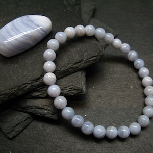 May include: A blue lace agate bracelet with matching tumbled stone. The bracelet features round, light blue beads. The tumbled stone is oval-shaped with a polished surface and a swirling pattern of blue and white. The items are displayed on a dark, textured surface.