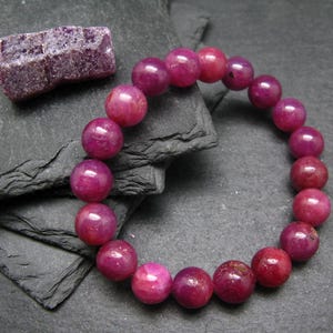Puede incluir: Una pulsera de cuentas con cuentas grandes y redondas de color rosa intenso. Una pequeña piedra púrpura en bruto se encuentra cerca. La pulsera se exhibe sobre una superficie oscura y texturizada, creando un contraste de colores y texturas.