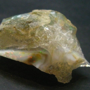 Może przedstawiać: Surowy, przejrzysty opal w jasnobrązowym kolorze z iryzującymi zielonymi, czerwonymi i białymi błyskami. Opal ma naturalny, nieoszlifowany i nieregularny kształt.