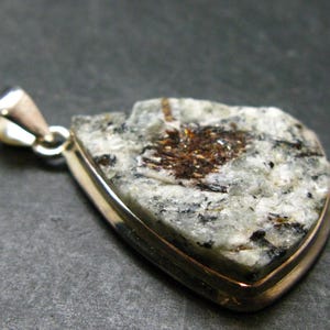 May include: A pendant featuring a rough, irregularly shaped stone with a speckled white, gray, and black surface, accented by a central dark brown crystalline inclusion. The stone is set in a smooth, polished gold-tone metal frame with a bail.