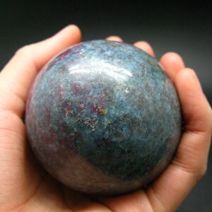 Puede incluir: Una esfera de piedra preciosa redonda y lisa, de color azul y rosa. La esfera está pulida y tiene un aspecto moteado.