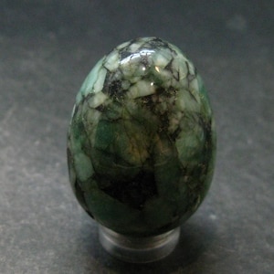 May include: A polished, egg-shaped gemstone featuring a mosaic-like pattern of deep forest green, translucent light green, and opaque black mineral inclusions throughout its smooth, rounded surface, resting on a small, clear circular base.