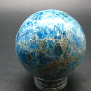 May include: A polished blue apatite sphere. The gemstone has a marbled pattern of deep blue and lighter blue hues, with hints of brown. The sphere is displayed on a clear stand, showcasing its natural beauty. A decorative item.