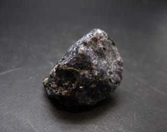 May include: A dark, rough-textured rock with a mix of dark gray, black, and hints of purple. The natural stone has an irregular shape and appears to be a mineral specimen. The lighting highlights the stone's surface details.