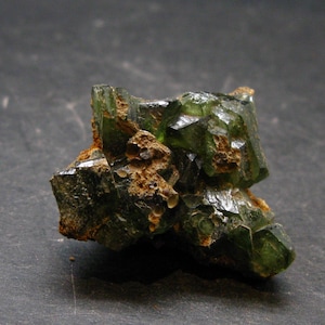 May include: A raw, uncut green crystal cluster with a rough, textured surface. The crystal has a dark green hue with areas of brown and tan. The facets of the crystal reflect light, creating a sparkling effect. The mineral specimen is set against a dark background.