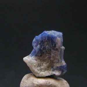 May include: A close-up of a raw, natural lapis lazuli gemstone. The stone features a deep blue color with flecks of white and gray. The gemstone is rough and irregular in shape, showcasing its natural beauty.