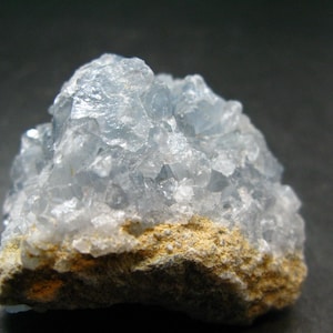 May include: A cluster of pale blue celestite crystals with a translucent, jagged appearance. The mineral formation features numerous small, sharp-edged crystalline points rising from a rough, tan-colored rocky base with a textured surface.