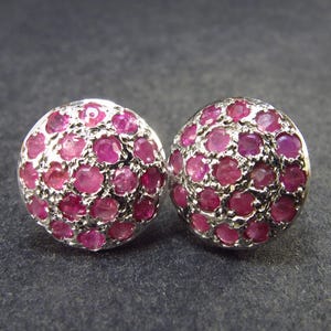 May include: A pair of silver stud earrings with pink ruby stones. The earrings are round and have a cluster of small stones.
