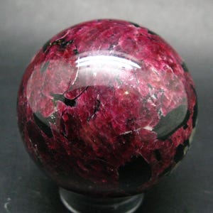 May include: A polished spherical stone featuring a vibrant deep magenta and dark red crystalline structure with contrasting black mineral inclusions throughout the surface, showcasing a smooth, reflective texture and natural internal fractures.