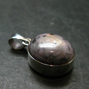 May include: A pendant featuring an oval-shaped, cabochon-cut star sapphire gemstone with a light purple hue and dark inclusions, set in a polished silver-toned metal bezel with a matching silver-toned metal bail for a necklace chain.