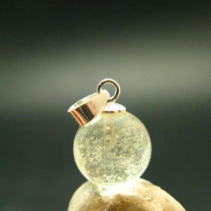 May include: A spherical pendant featuring a clear, textured glass orb with internal inclusions, topped with a polished silver-toned metal bail. The pendant has a smooth, rounded surface and a reflective metallic attachment for a chain or cord.